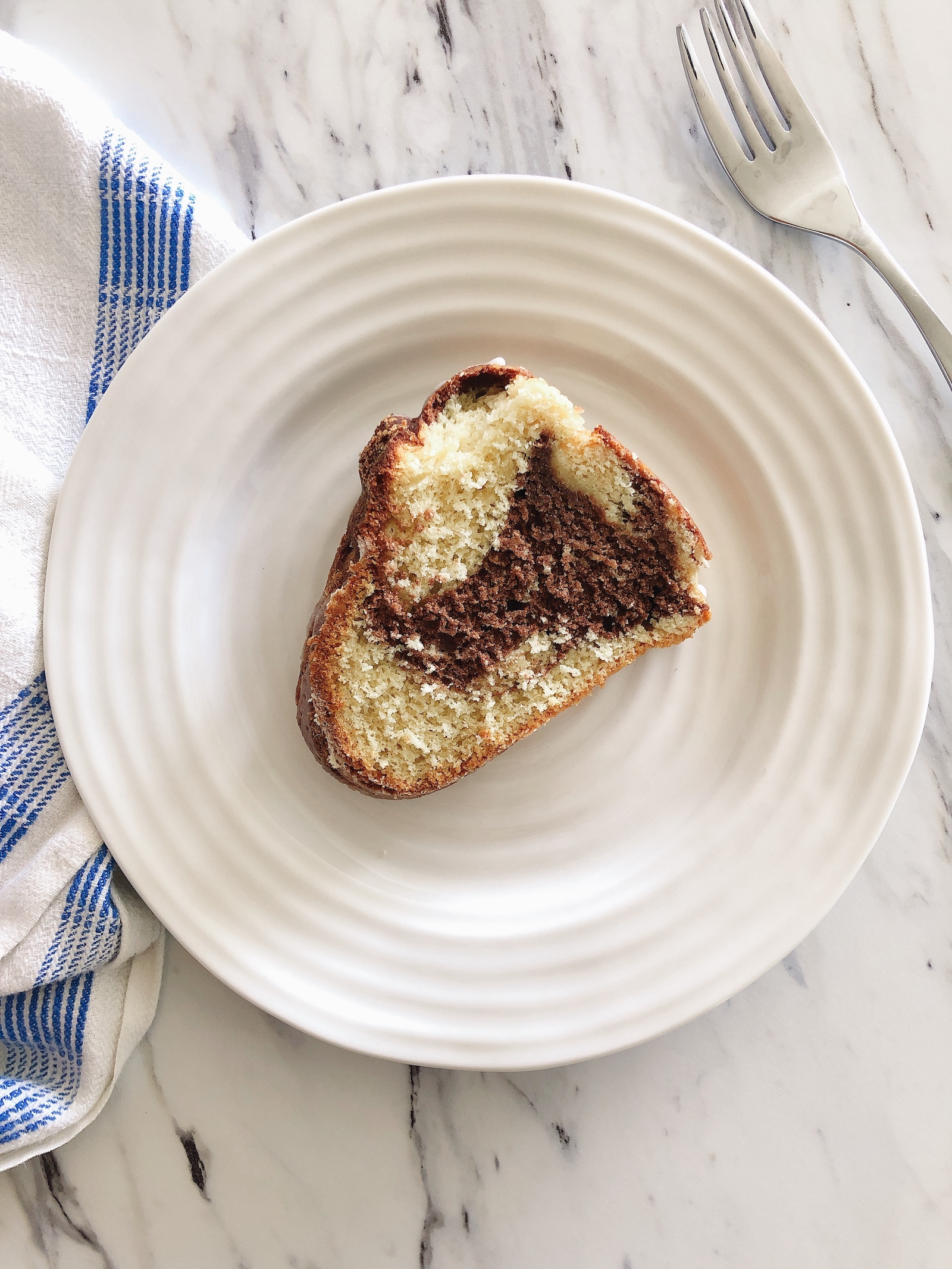One slice of vanilla and chocolate spelt marble cake lying on its side on a small white plate with a blue and white tea towel tucked under the plate.