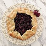 A rustic cherry galette on a decorative pie serving plate with three fresh cherries on the side