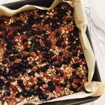 A square baking pan lined with parchment paper and filled with ancient grain blueberry banana baked oatmeal studded with toasted pecans