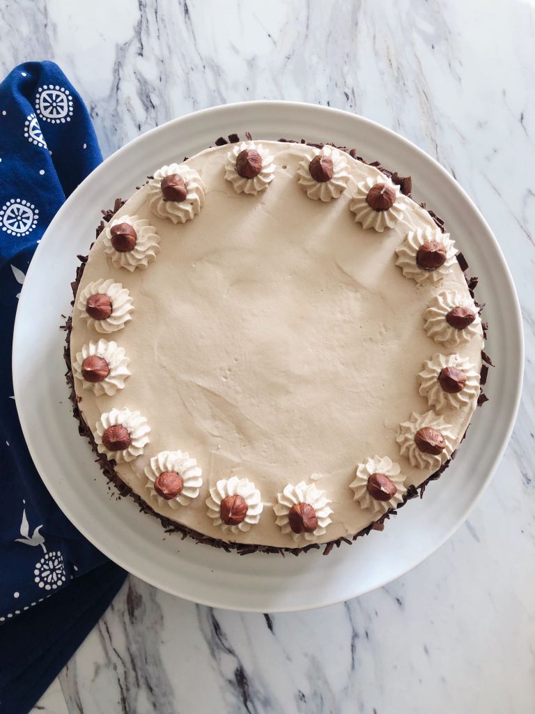A Hazelnut Mocha Spelt Torte decorated with twelve whole hazelnuts and chocolate shavings. The torte is on a white cake plate with a blue tea towel and white marble countertop in the background