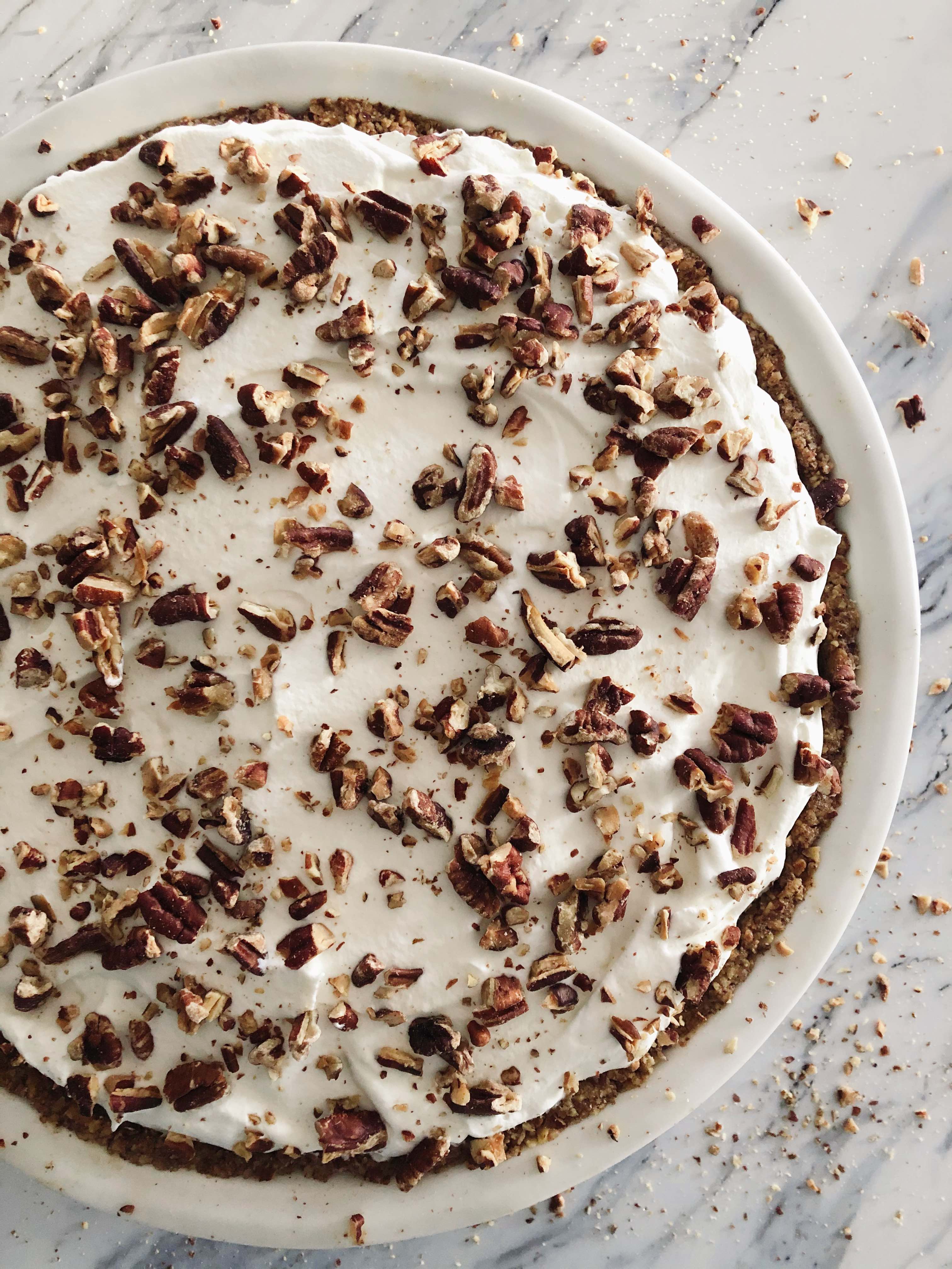 Pumpkin Pecan Torte topped with whipped cream and chopped pecans in a white pie plate on a white marble countertop