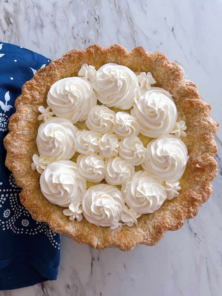 Banana Cream Pie with swirls of whipping cream on top of various sizes. A blue with white pattern tea towel lays underneath.
