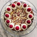A round white chocolate cheesecake with raspberry swirl on a glass cake plate topped with twelve whipping cream swirls and a raspberry on top of each swirl