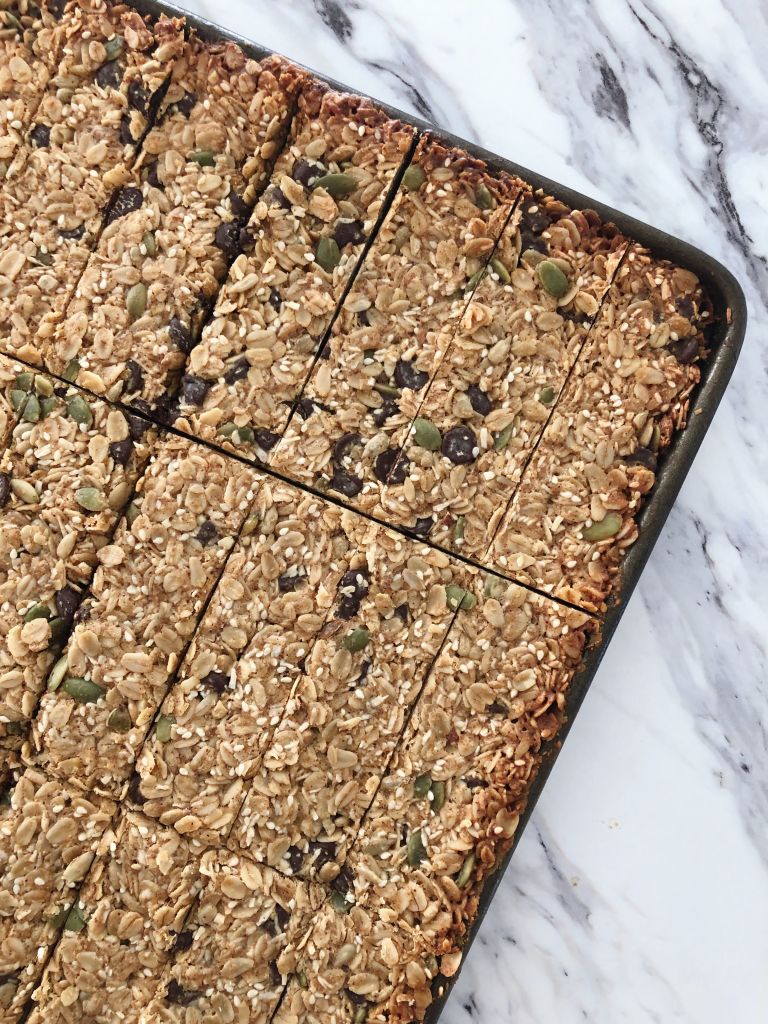 A baking sheet pan full of Homemade Chocolate Chip Granola Bars with Pumpkin, Sunflower and Sesame Seeds cut into thin rectangles on a white marble countertop.