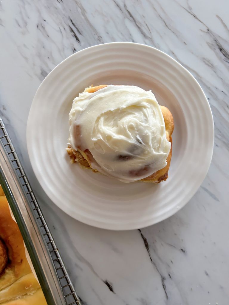 One fluffy sourdough discard cinnamon bun covered with cream cheese icing sitting on a small round white plate. 