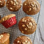Six rhubarb and applesauce whole grain spelt muffins cooling on a wire rack. Two of the muffins are lying on their side to show the red and yellow muffins cups they were baked in.